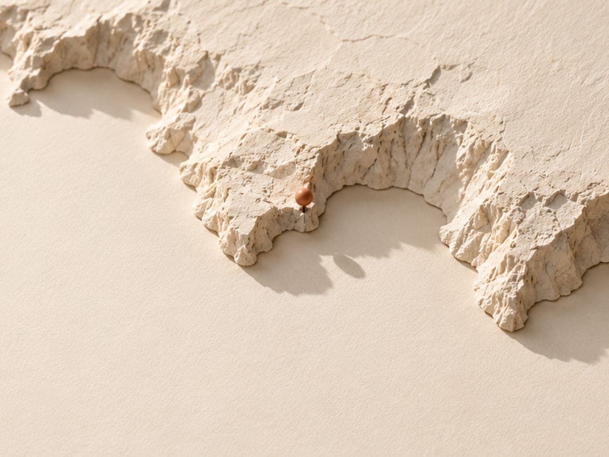 Une carte en relief de papier ivoire dessinant des calanques, une épingle terre cuite plantée au bord d’une crique.